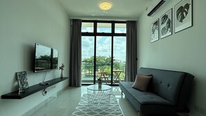A modern living room with a flat-screen TV, a grey sofa, a coffee table, and a view of greenery through large windows. - Green Haven Comfort House (Masai)
