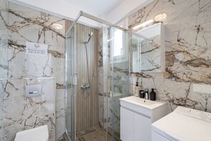 A modern bathroom with a glass-enclosed shower, marble walls, a white sink, and a built-in washing machine.