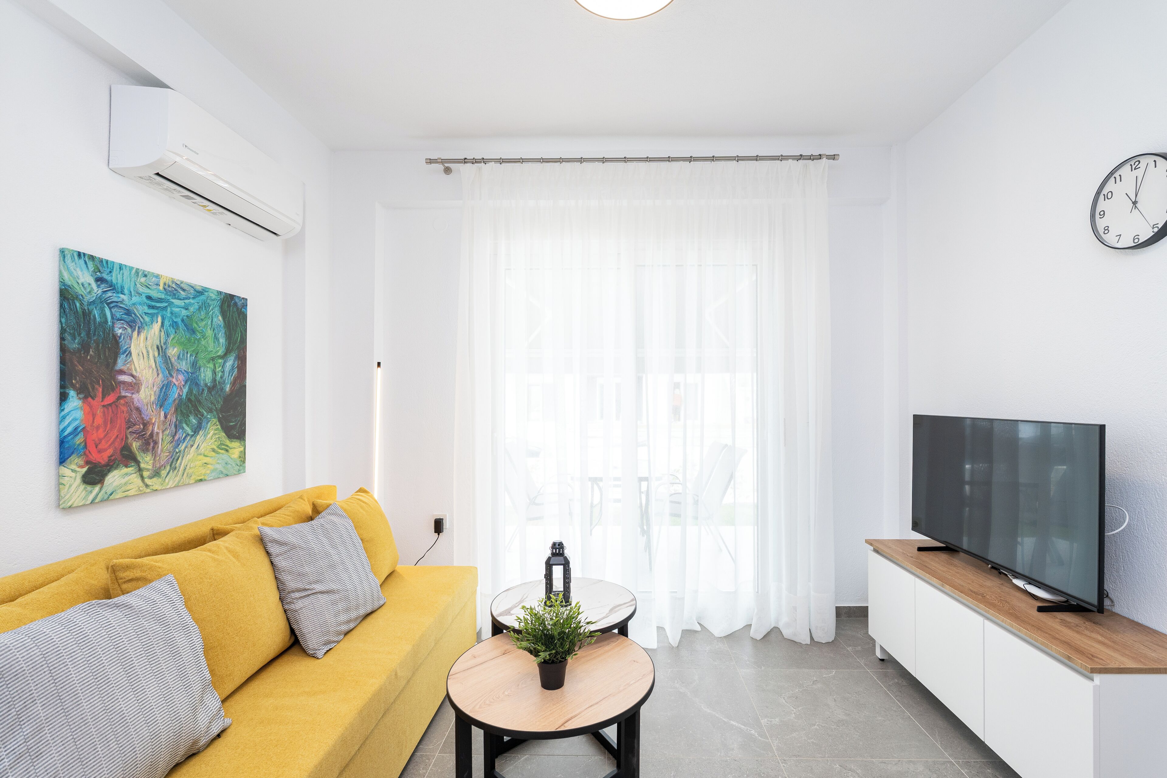 Un saló modern amb un sofà groc, una taula rodona petita amb una planta, un televisor de pantalla plana sobre un moble de fusta i una finestra gran amb cortines lleugeres.