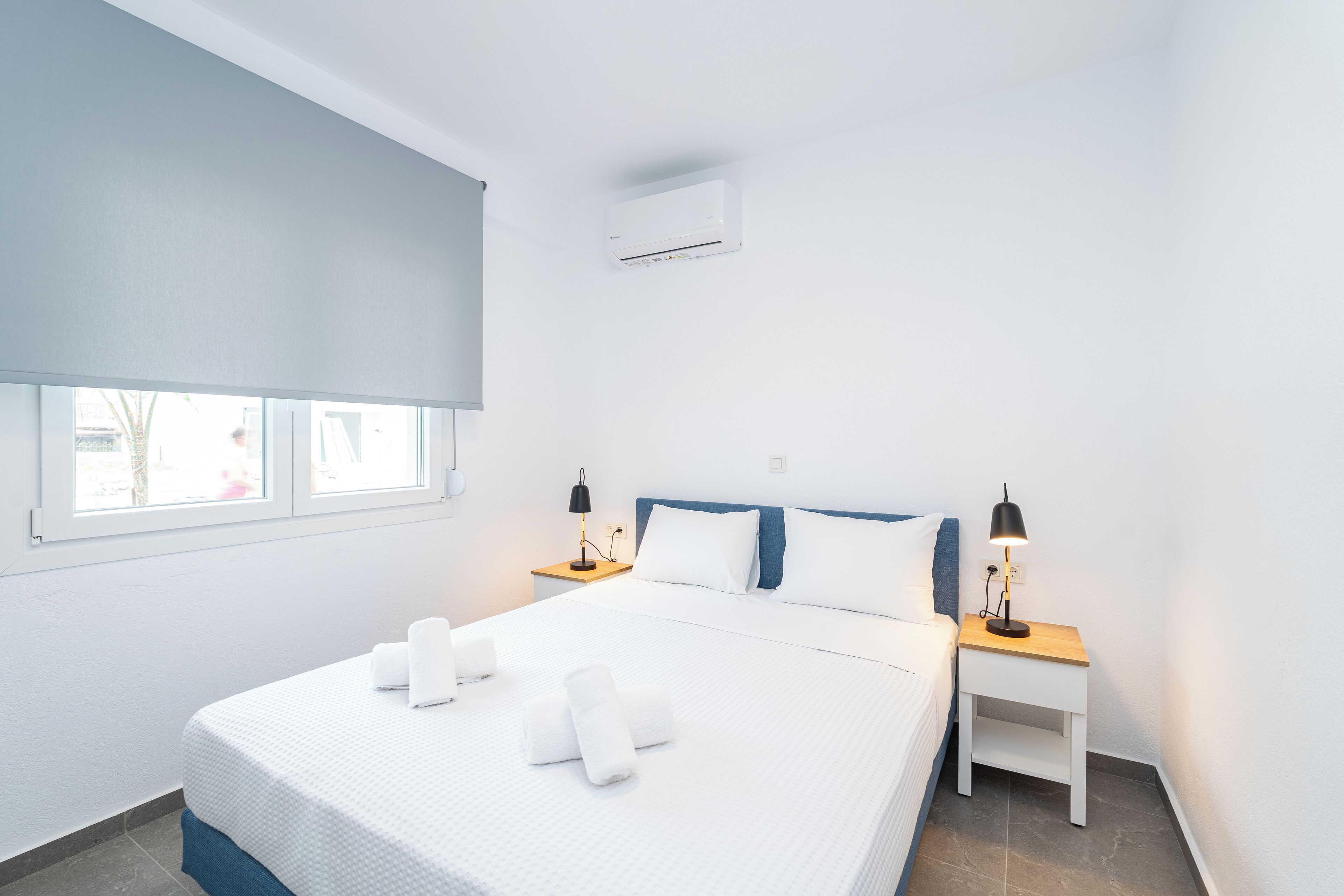 A neatly arranged hotel room with a bed, two bedside tables with lamps, a window with a blind, and an air conditioning unit.