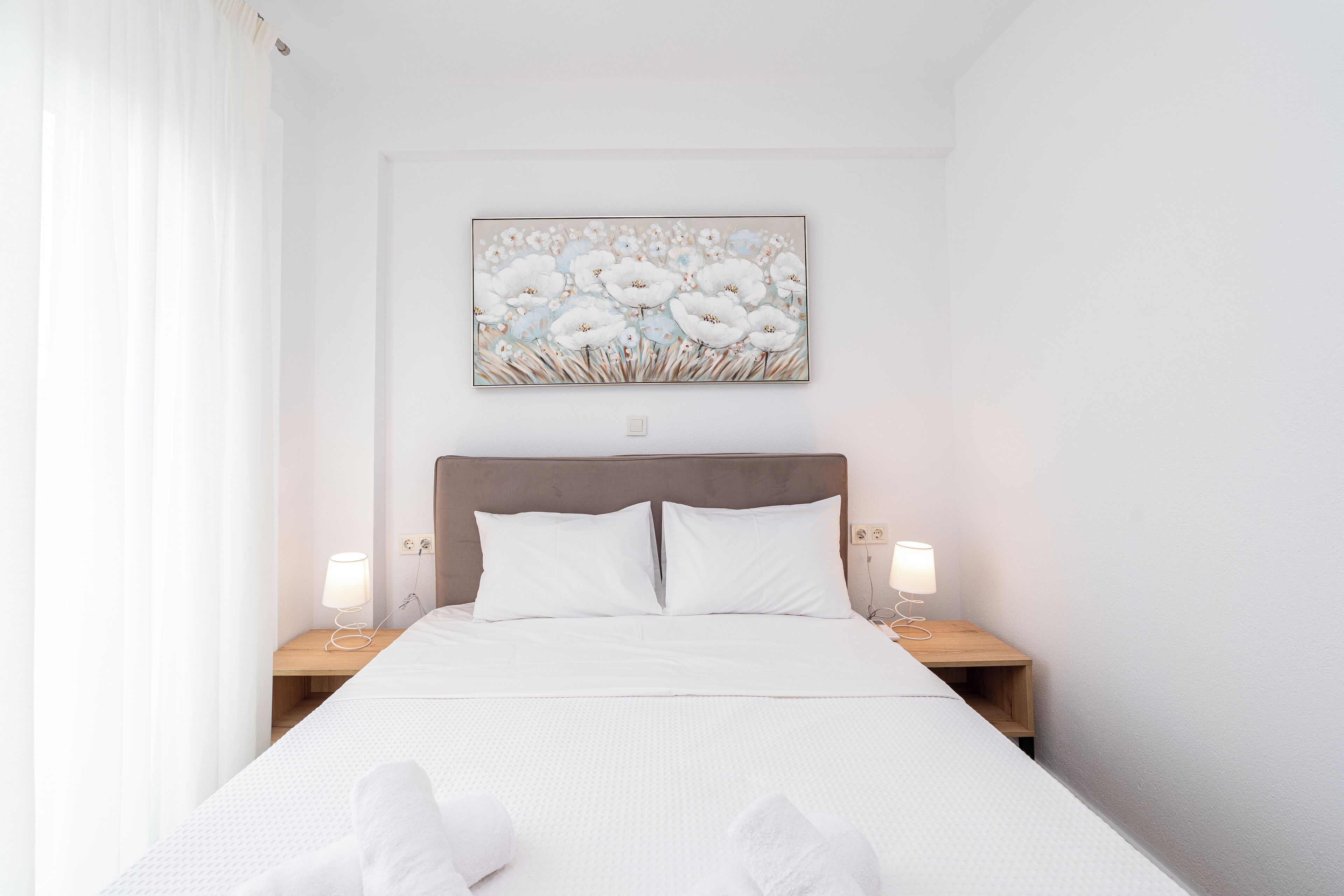 A neatly made bed with white linens, two bedside tables with lamps, and a wall-mounted painting of flowers.