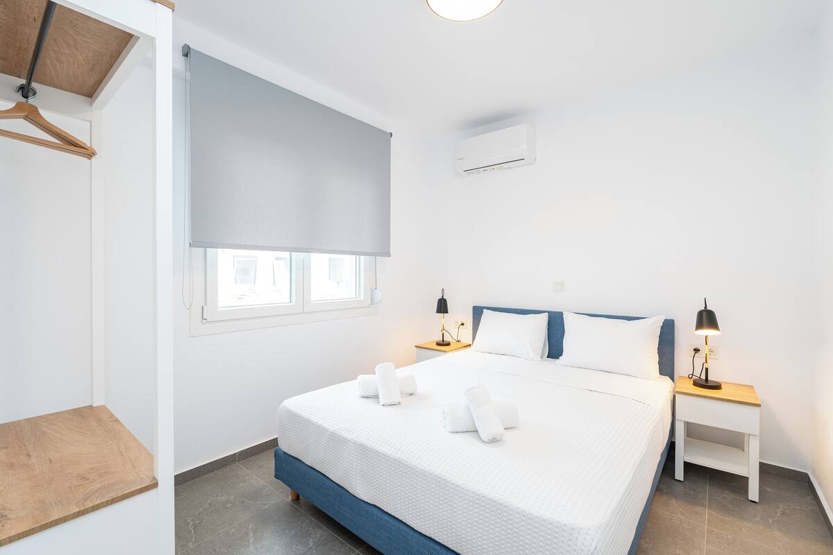 Ein modernes Hotelzimmer mit einem großen Bett, zwei Nachttischen, einer Holzablage, einem Fenster mit Rollos und einer Klimaanlage.