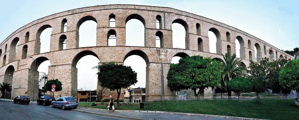 Une grande structure en pierre à arcades, dotée de plusieurs ouvertures, entourée d’une zone herbeuse et d’arbres.