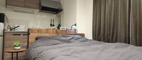 Habitación de hotel compacta con cama, una mesita con planta, kitchenette y un ventanal con cortinas.