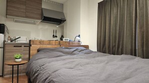 A compact hotel room with a bed, a small table with a plant, a kitchenette, and a large window with curtains.