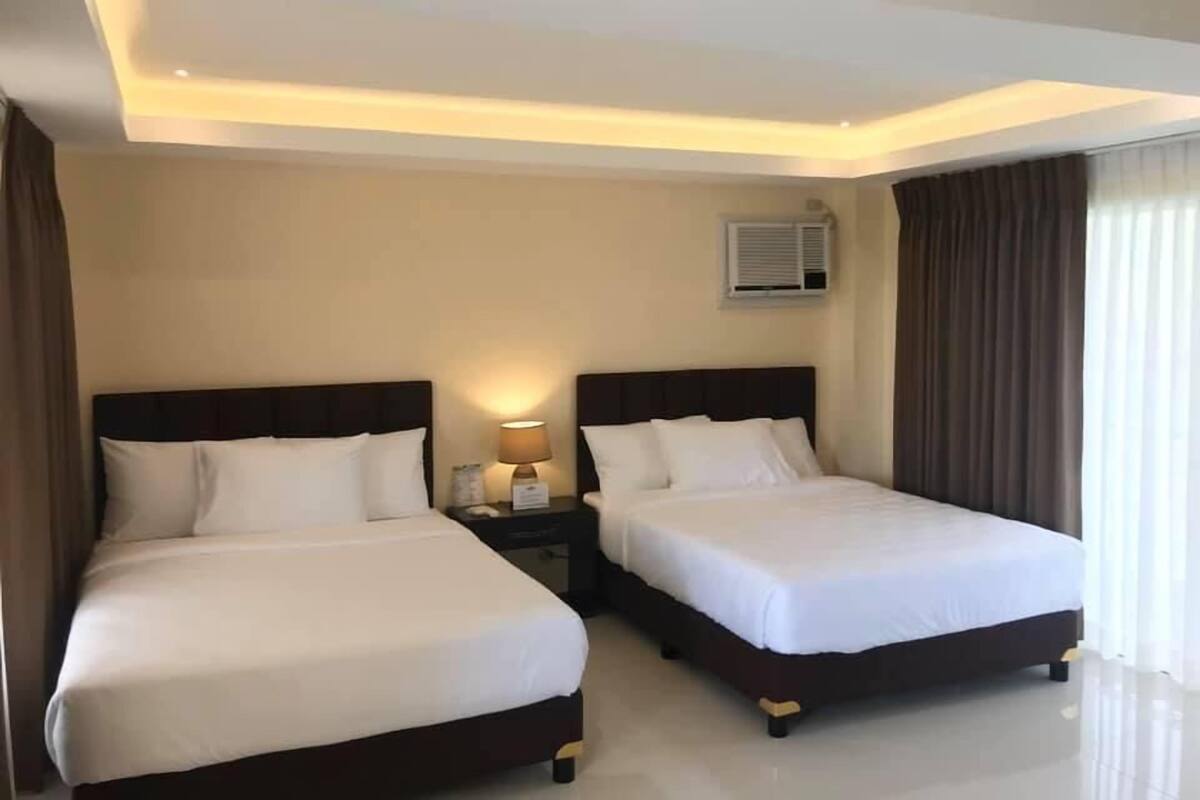 A hotel room with two beds, a nightstand with a lamp, an air conditioning unit, and a window with curtains.