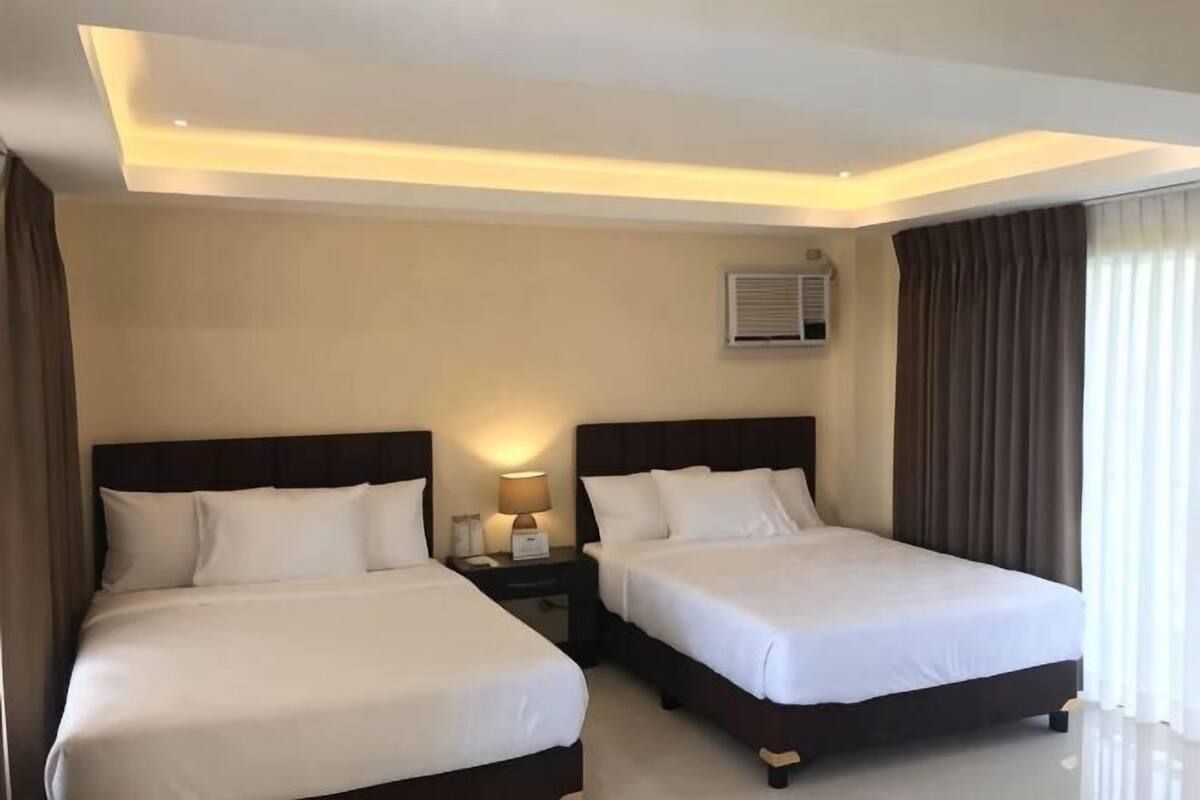 A hotel room with two beds, a nightstand with a lamp, a ceiling light, and an air conditioning unit.