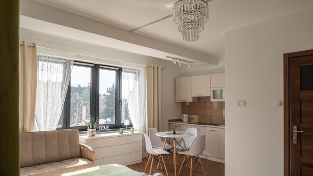 A compact living space with a sofa, a dining table with chairs, a kitchenette, and a large window with curtains. - Przytulny apartament KOLEJOVA 13 (Nowy Targ)
