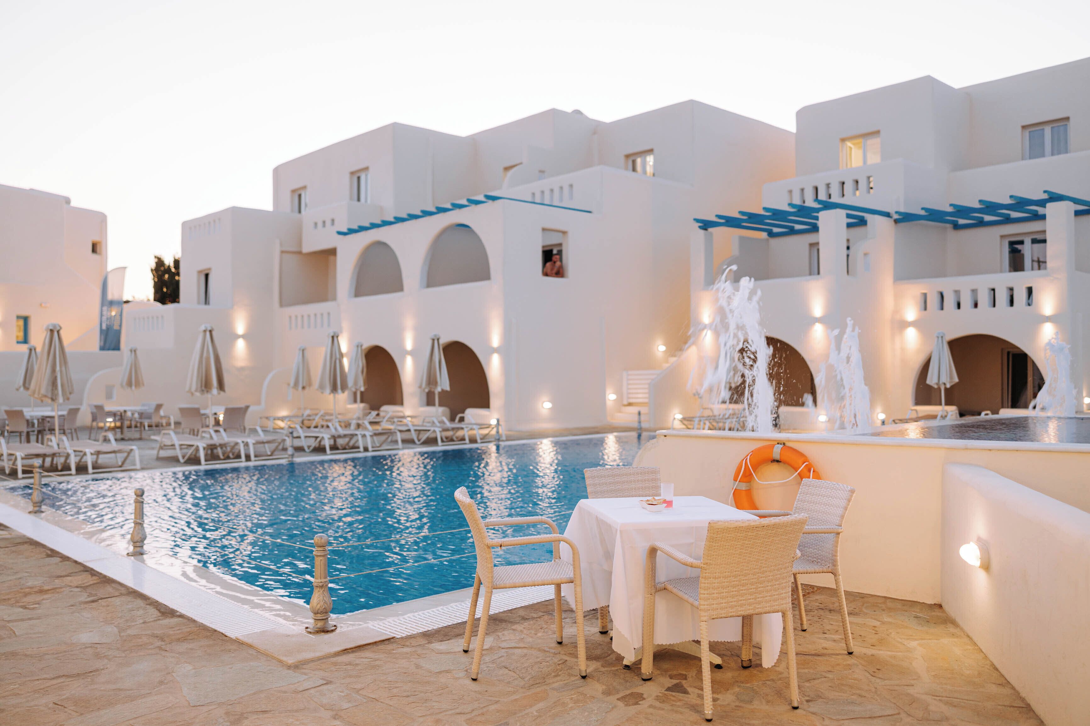An indoor swimming pool with lounge chairs and tables, surrounded by white buildings with blue accents.