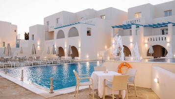 An indoor swimming pool with lounge chairs and tables, surrounded by white buildings with blue accents.