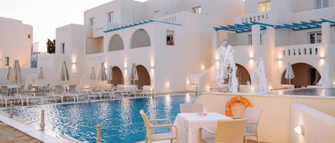 An indoor swimming pool with lounge chairs and tables, surrounded by white buildings with blue accents.