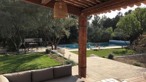 Ein überdachter Patio-Bereich mit Holztisch und einer Couch, im Hintergrund ein Pool und ein gepflegter Rasen.