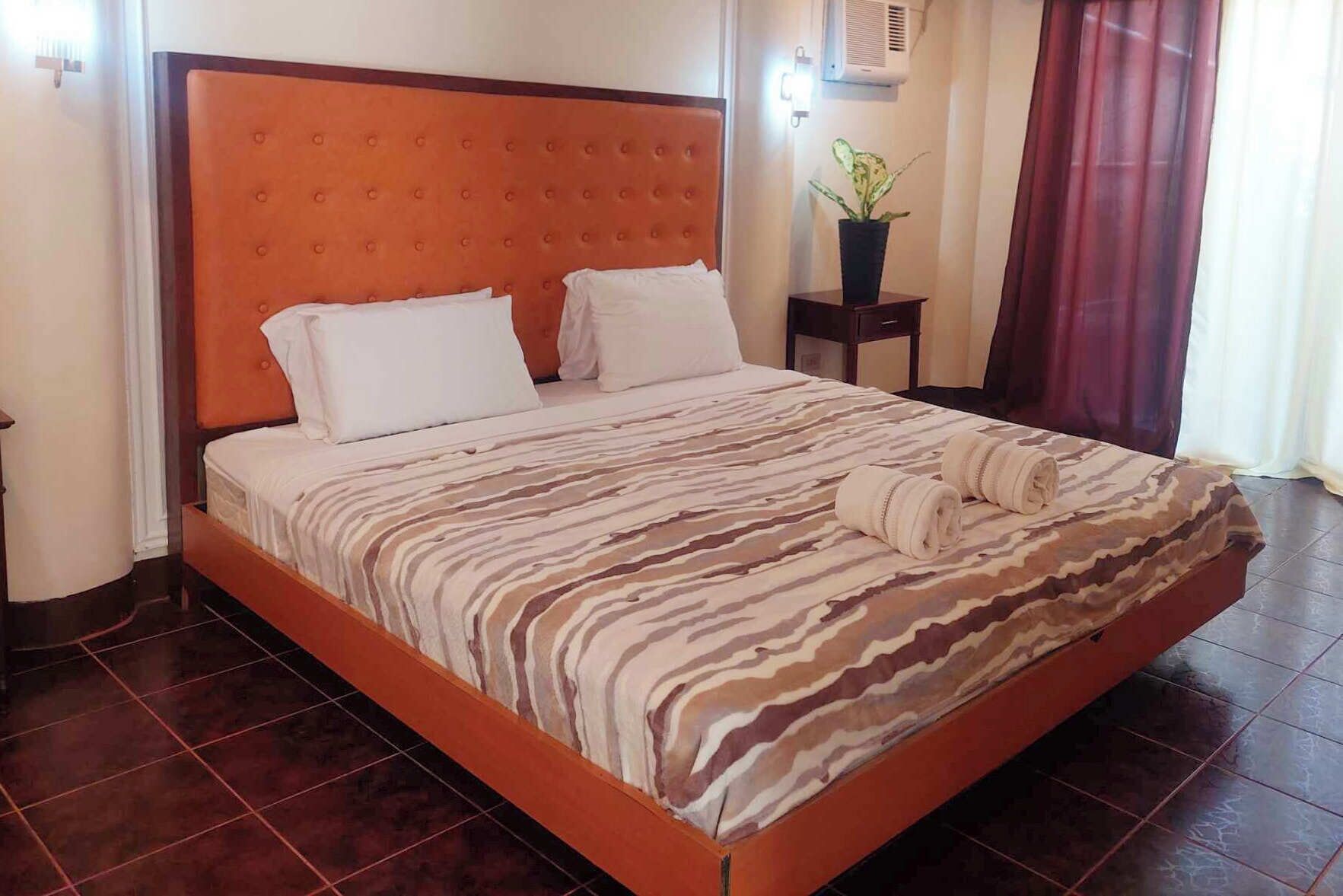 A hotel room with a large bed, a headboard, a bedside table with a plant, and a window with curtains.