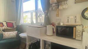 A compact kitchen with a microwave, toaster, and kettle on a white countertop. A window with curtains allows natural light, and a green chair is visible. - Cosy Irish Cabin studio in fabulous Derry with WiFi & Private Bathroom (Derry)
