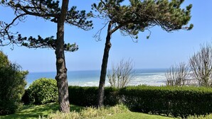 A coastal landscape with two prominent pine trees, a hedge, and a view of the ocean. - Le Nid des Falaises, au bord de la plage (Varengeville-sur-Mer)