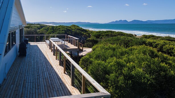 A wooden deck with outdoor seating, overlooking a coastal landscape with mountains in the distance. - Nine Mile Beach House - Holiday Houses Tasmania (Glamorgan-Spring Bay)