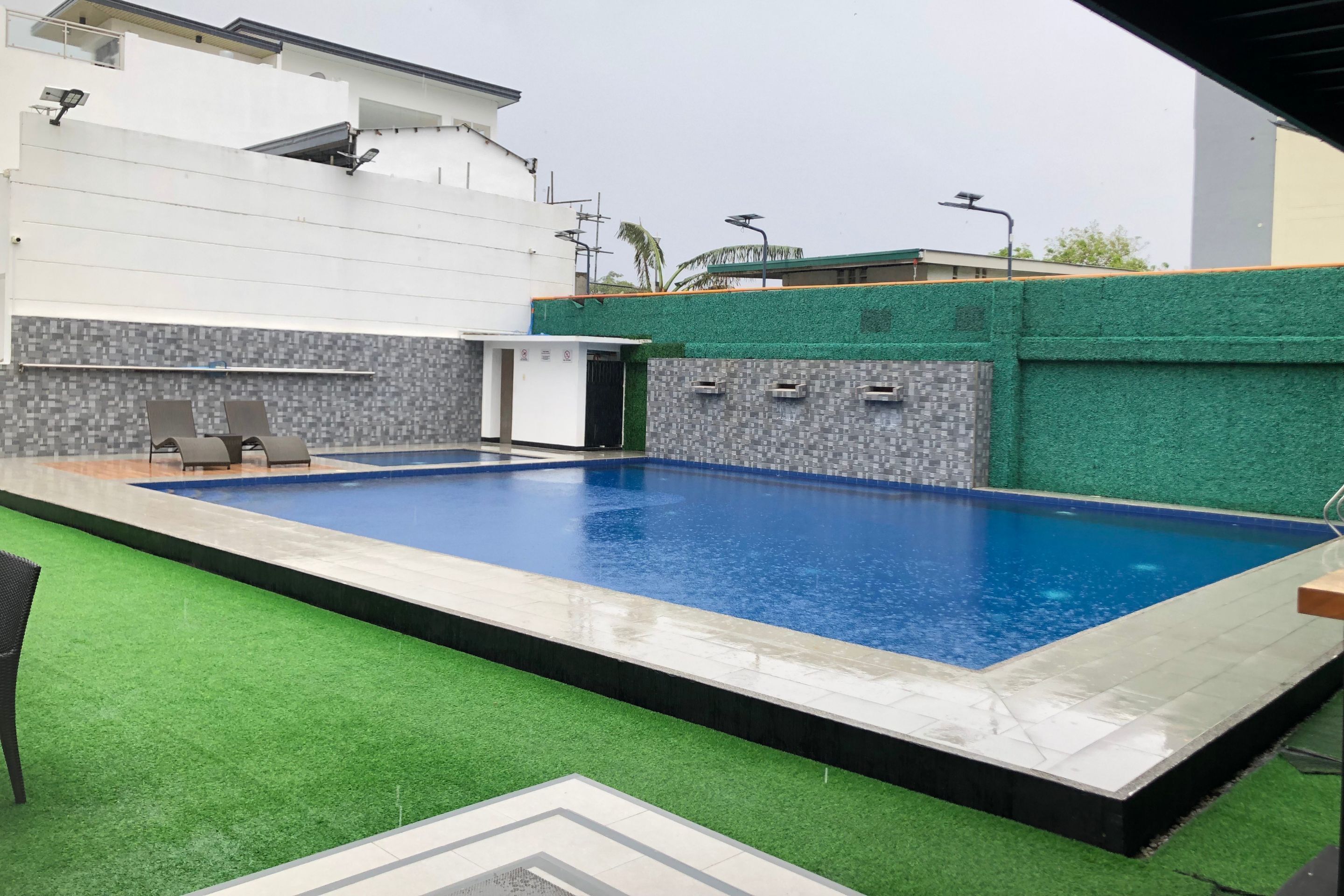 Una piscina rectangular con borde de azulejos, rodeada de césped artificial verde y un muro de azulejos con un diseño gris.