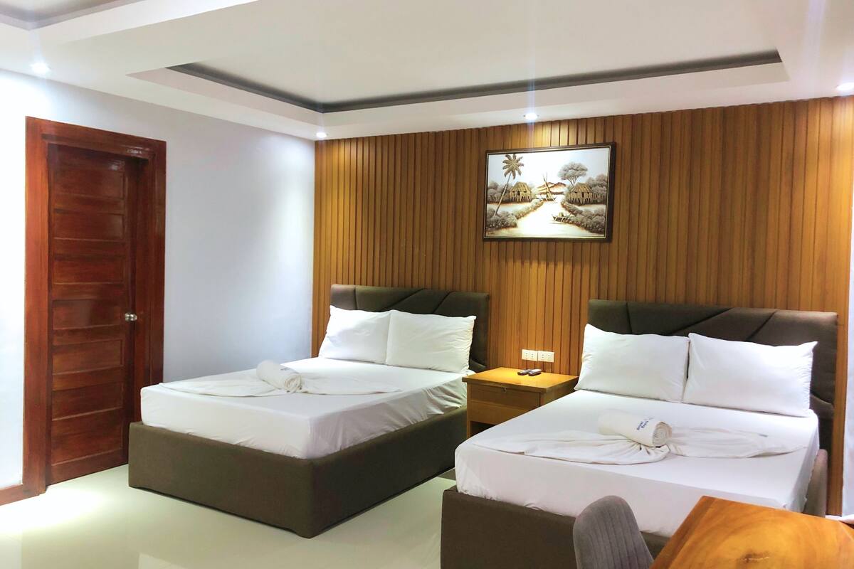 Quarto de hotel com duas camas, painéis de madeira nas paredes, um quadro e uma porta de madeira.