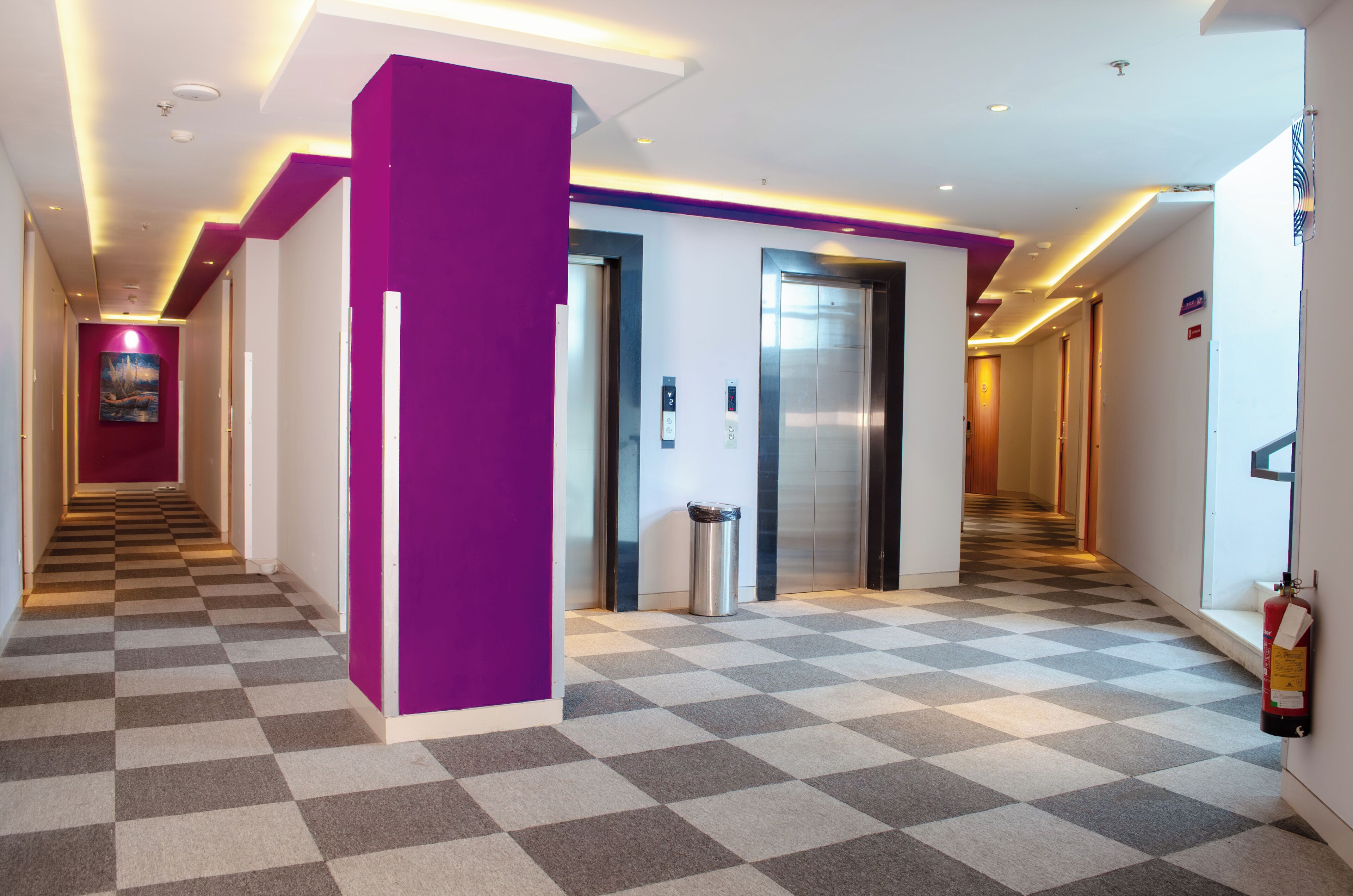 A hotel corridor with a checkered floor, elevators, a trash bin, and a fire extinguisher.