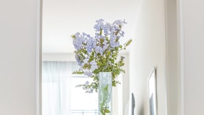A wooden console table with a white towel and a wicker basket below it, a blue bottle of reed diffuser, and a vase with flowers on the table.