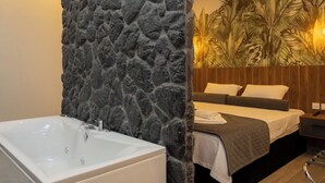 A bathroom with a stone wall, a bathtub, and a bed with a wooden headboard and a tropical wallpaper design. - Dioroh Suites Meteora 302 (Kalampaka)