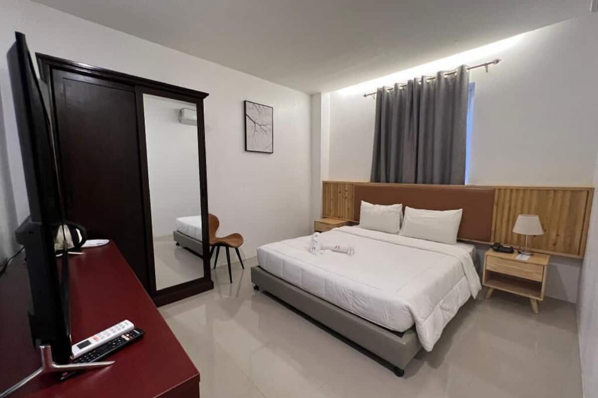 A hotel room with a bed, a desk, a chair, a nightstand, a lamp, a mirror, and a window with curtains.