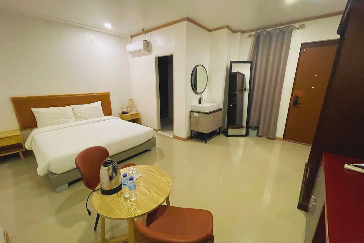 A hotel room with a bed, a small table with a kettle and water bottles, a chair, a desk with a mirror, and a bathroom with a sink and toilet.