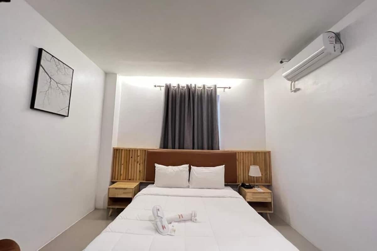 A hotel room with a bed, a headboard, a nightstand, a lamp, a wall-mounted air conditioner, and a window with curtains.