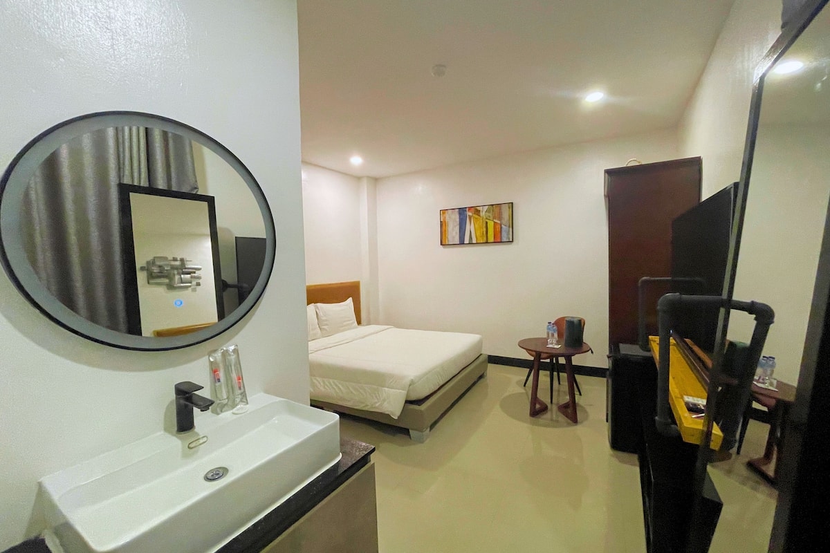 A hotel room with a bed, a round mirror, a sink, a small table, and a TV.