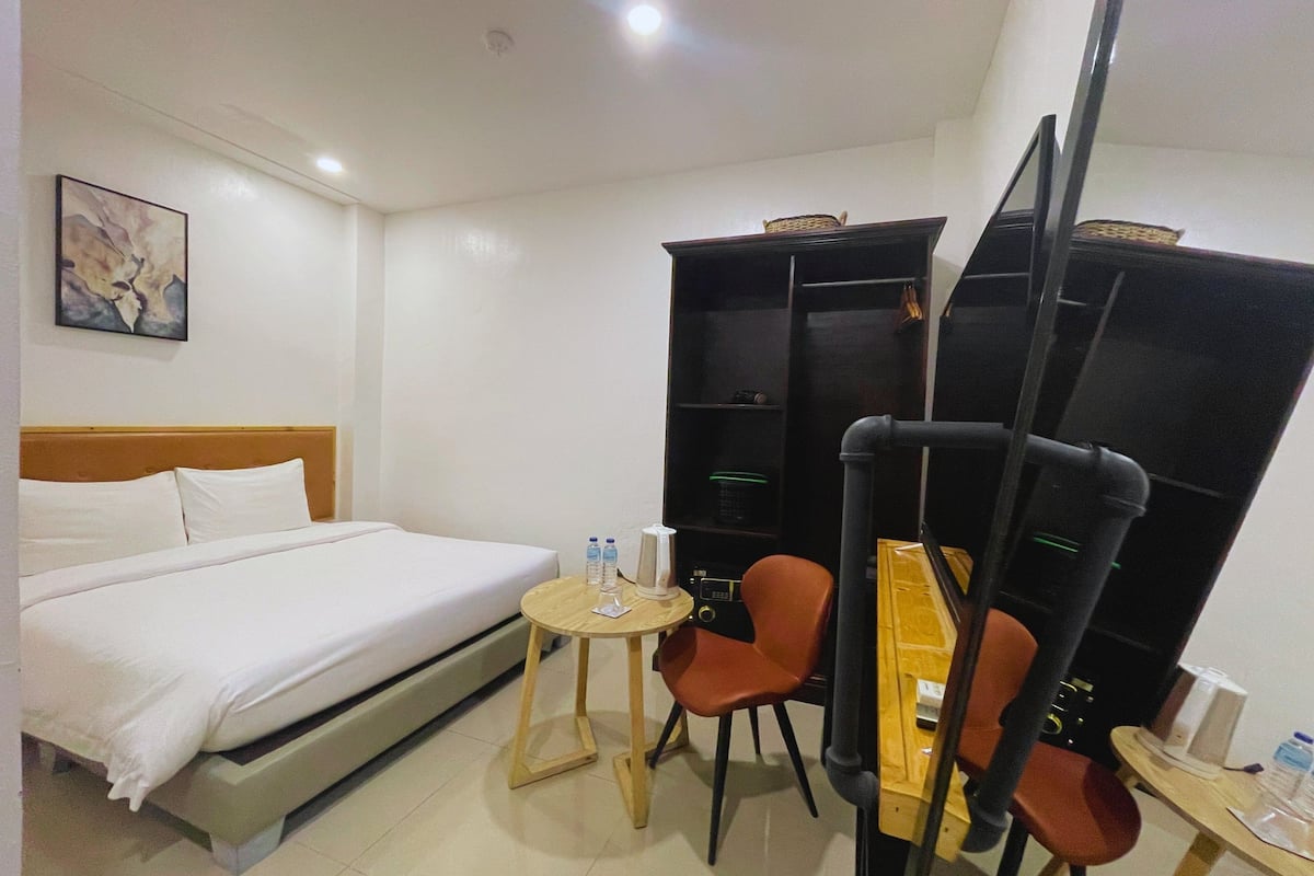 A hotel room with a bed, a small round table with two bottles, a chair, and a bookshelf.