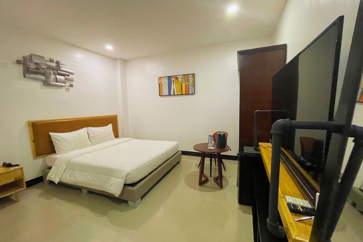 A hotel room with a bed, a small table with a chair, a TV, and a painting on the wall.