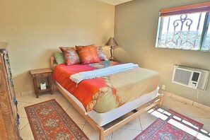 Ein Schlafzimmer mit Bett, Nachttisch, Lampe, Fenster, Klimaanlage und gemusterten Teppichen.