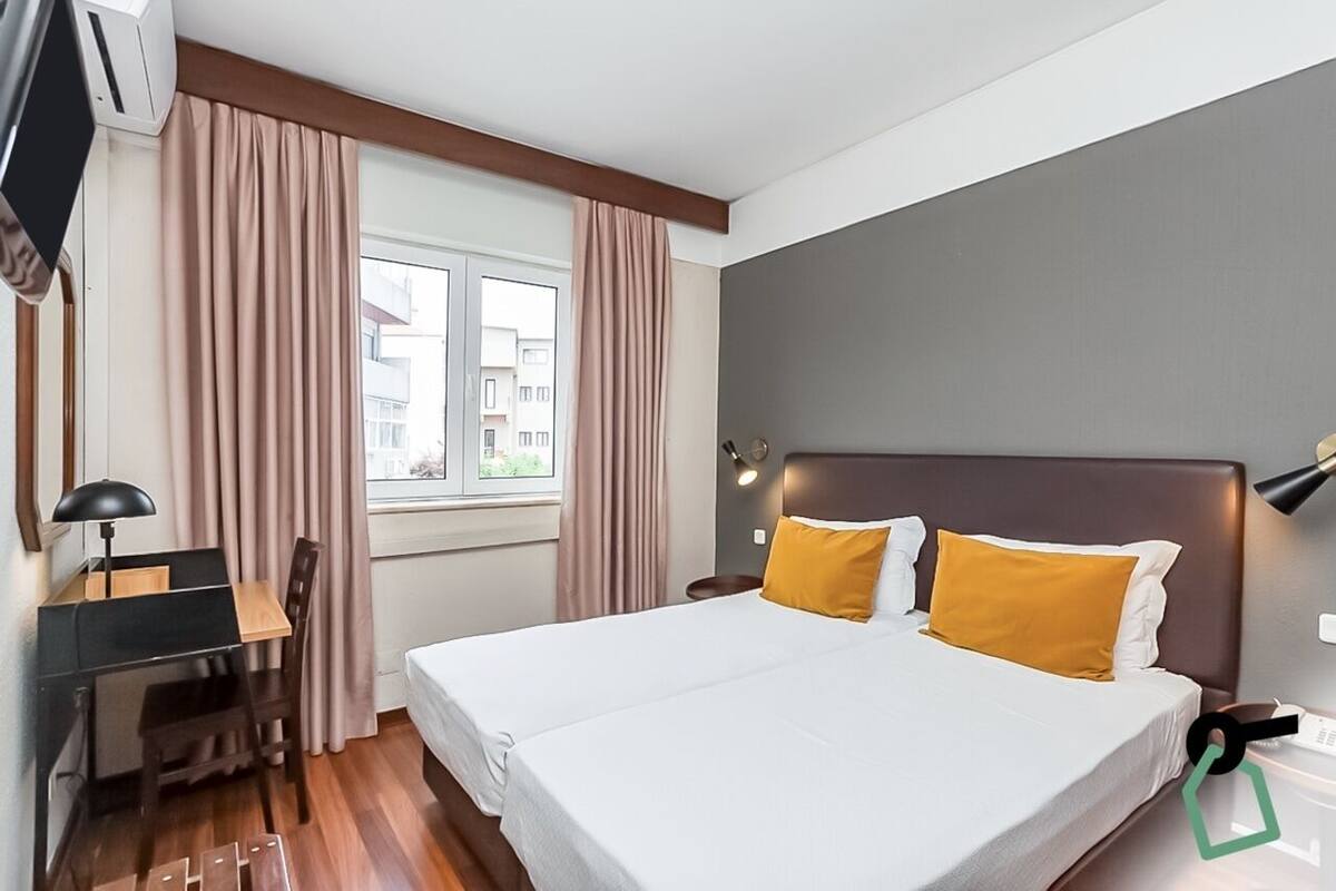 A hotel room with a bed, a desk, a chair, a window with curtains, and a wall-mounted lamp.