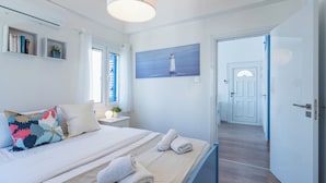 Une chambre à coucher avec un lit, des oreillers, un tableau représentant un phare, une table de chevet avec une plante, et une porte donnant sur une autre pièce.