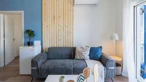 Un salon moderne avec un canapé gris, une petite table d’appoint ornée d’une plante, une table basse blanche et une grande fenêtre avec des stores.