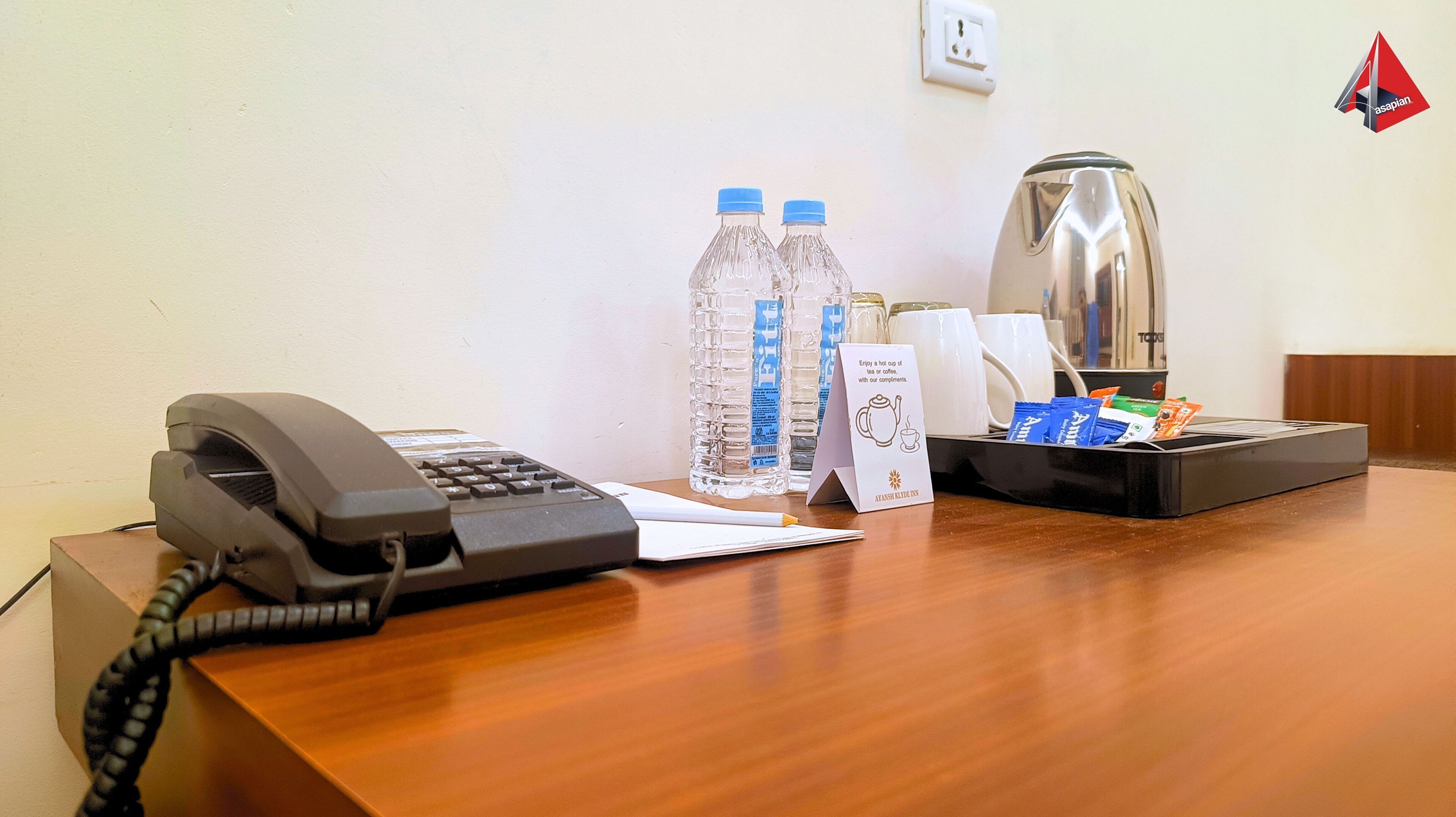 A hotel desk with a telephone, two water bottles, a coffee maker, and a tray with coffee cups and snacks.
