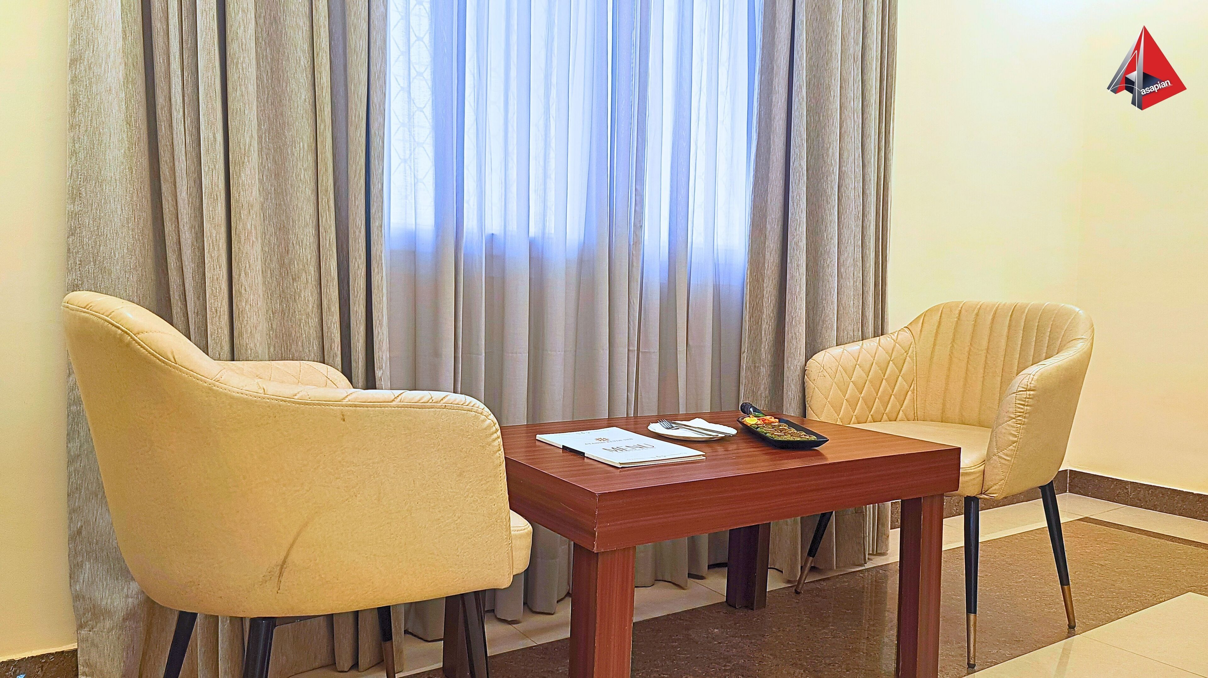A hotel room with two beige armchairs, a wooden table, and a window with grey curtains.