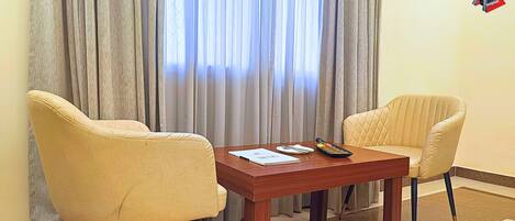 A hotel room with two beige armchairs, a wooden table, and a window with grey curtains.