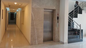 A modern hotel corridor with a marble floor, a central elevator, and a staircase with metal railings. - Hotel The Luxem (Gurugram)