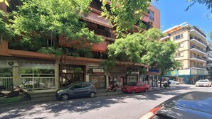 Une vue de rue avec des voitures garées, un immeuble à plusieurs étages et des arbres qui offrent de l’ombre.
