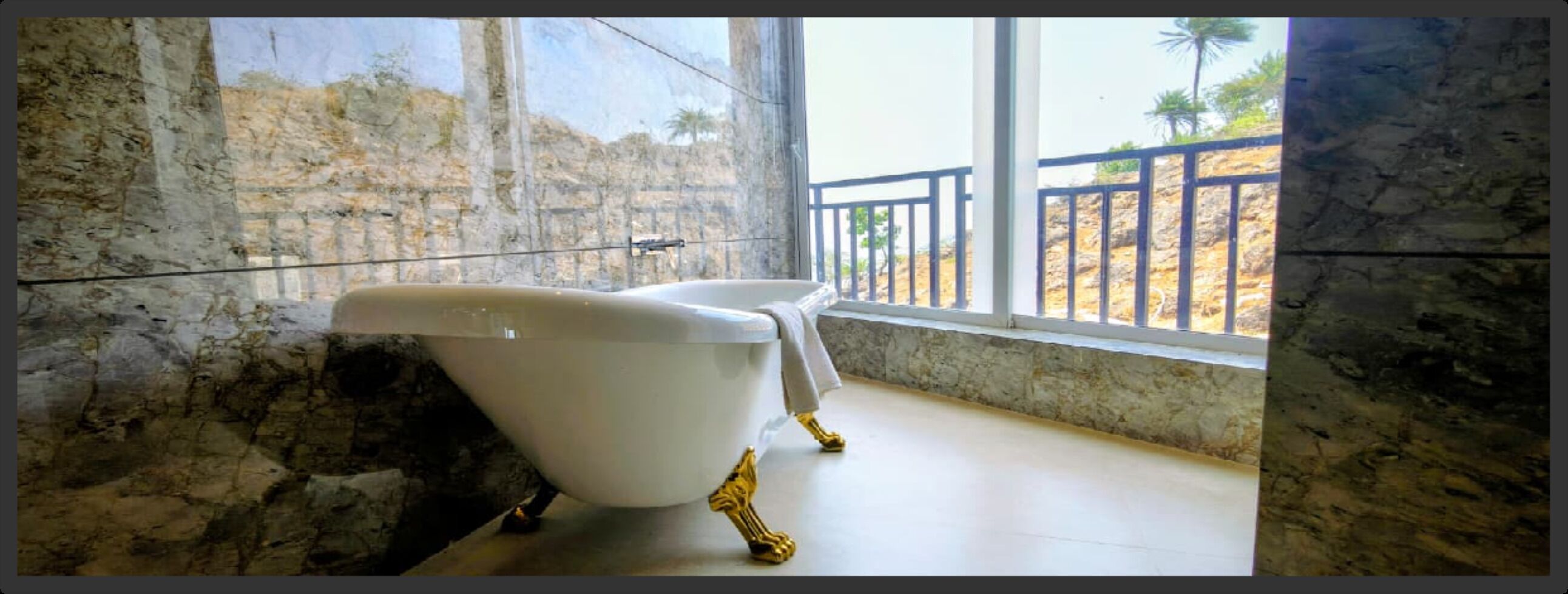 A bathtub with golden legs in a room with stone walls and a view of palm trees.