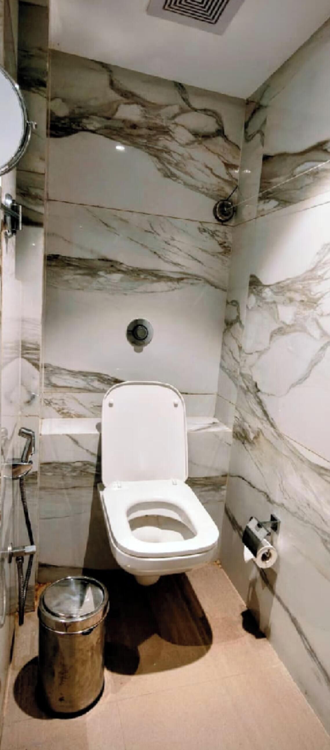 A bathroom with a toilet, marble walls, a metal trash bin, and a shower area.