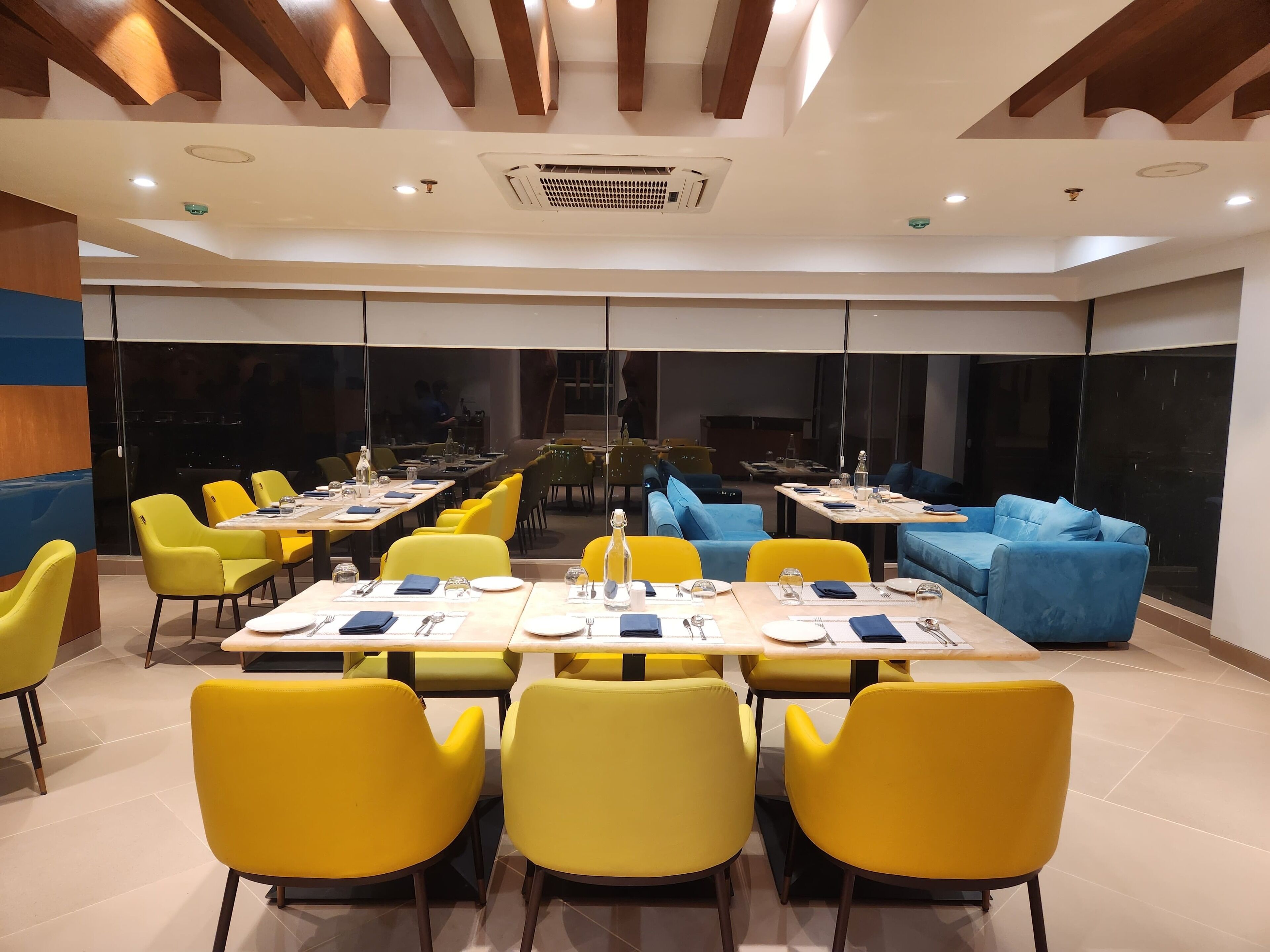 A modern dining area with yellow chairs, tables set for dining, and a blue sofa in the background.