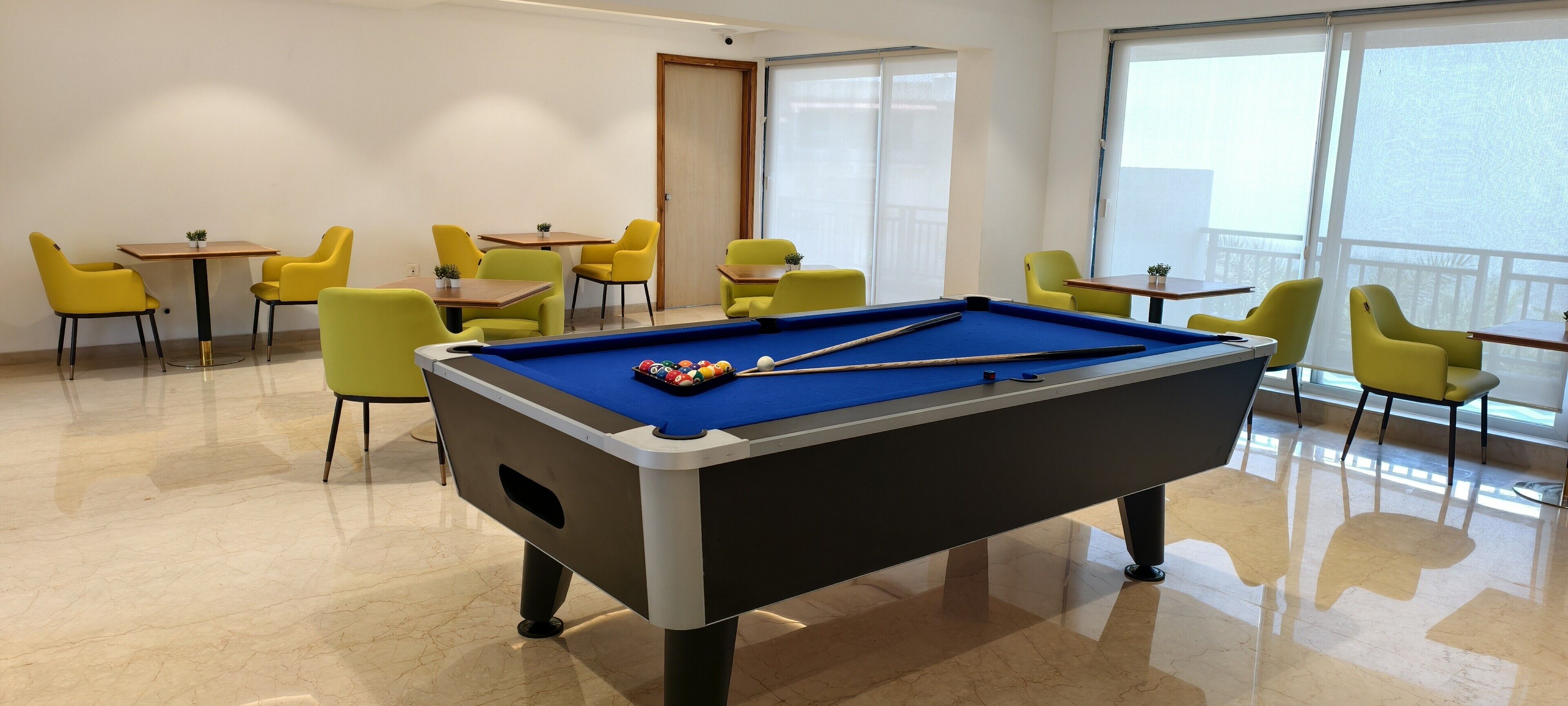A pool table with billiard balls in a room with yellow chairs and tables, large windows, and a door.