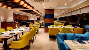 Interior de un restaurante moderno con asientos amarillos y azules, una zona de cocina central y un diseño de techo contemporáneo.