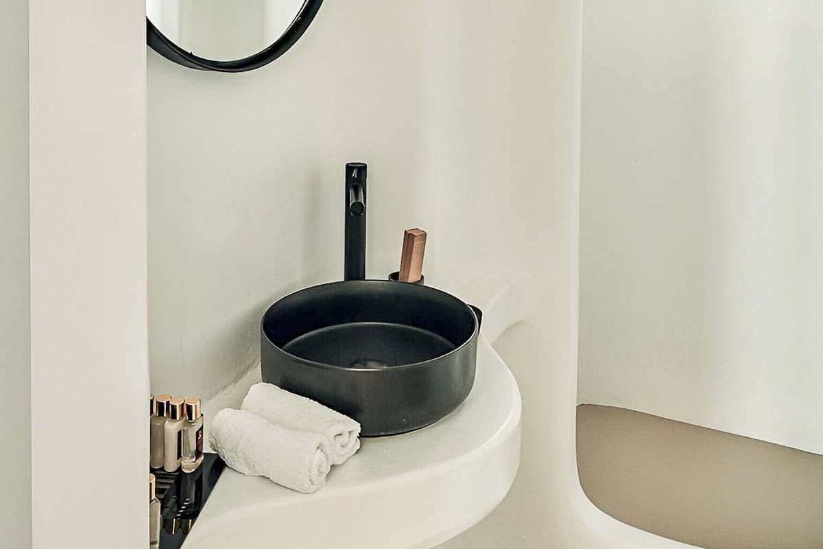 Une salle de bain avec un miroir rond, un lavabo noir, une fenêtre et un porte-serviettes.