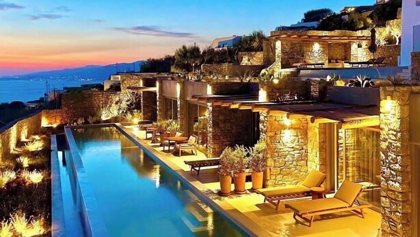 A poolside area with stone walls, lounge chairs, and a view of the sea at dusk. - Hotiday Mykonos Fanari (Mykonos)