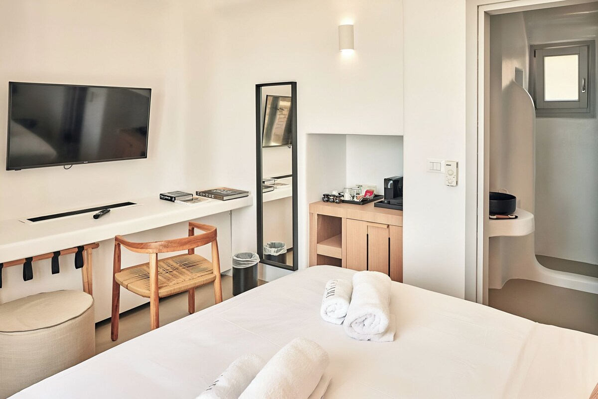 A hotel room with a bed, a desk with a TV, a chair, a mirror, a bathroom with a sink, and a door.