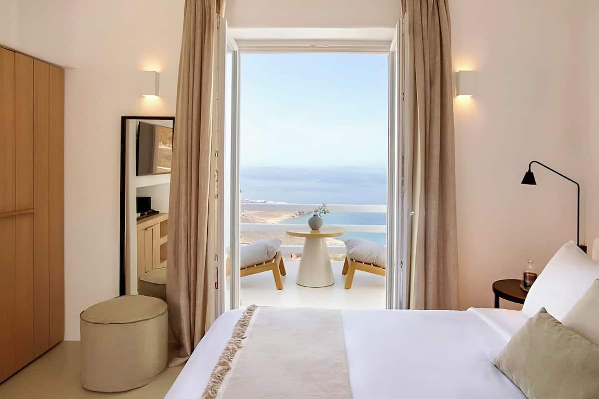 A bedroom with a large bed, a view of the sea, and a balcony with a table and chairs.