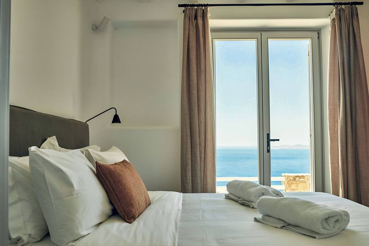 A hotel room with a bed, a nightstand, a lamp, a window with curtains, and a view of the sea.